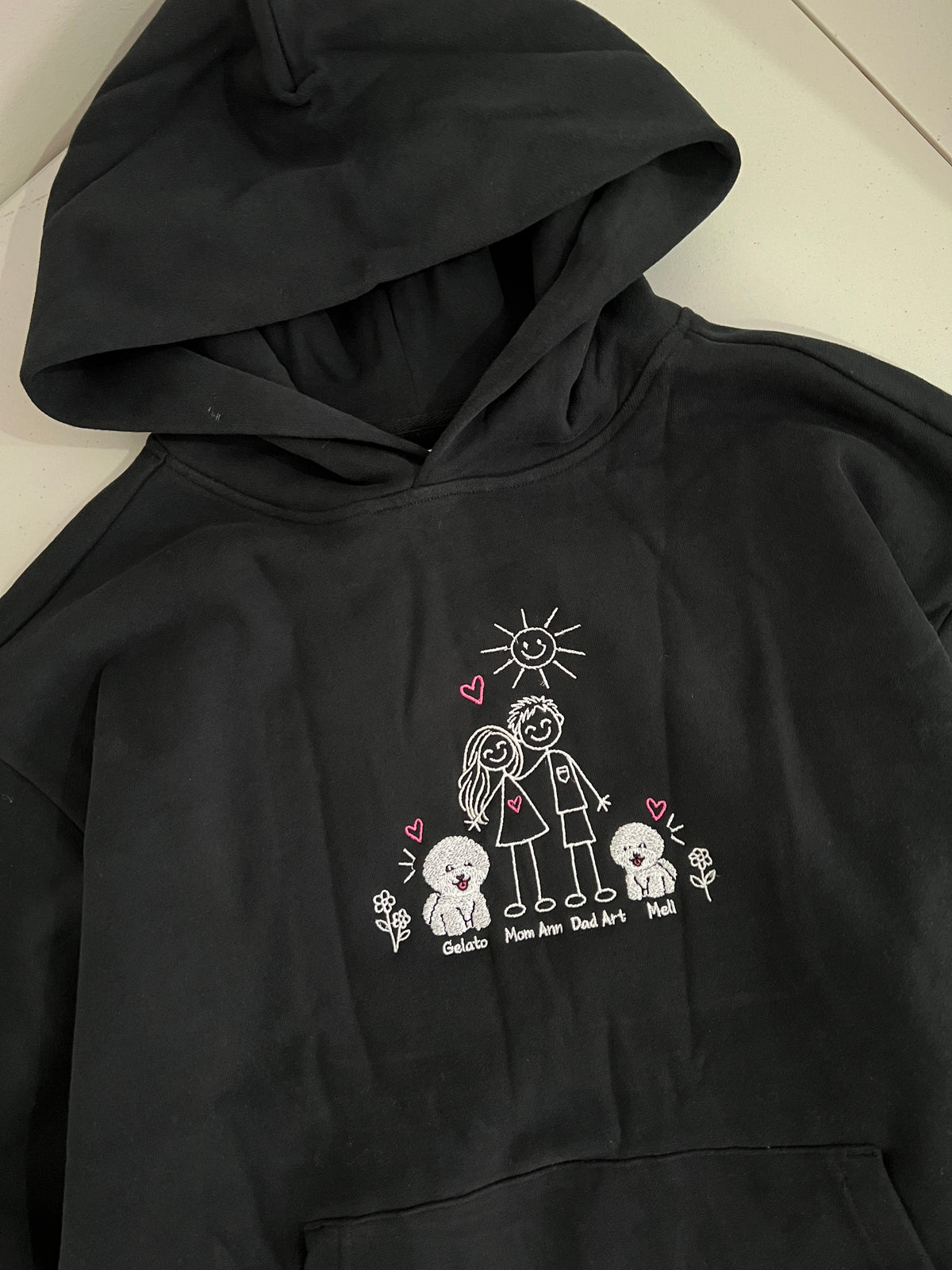 A black pullover hoodie with a white hand-drawn style graphic on the front showing a couple, a sun, hearts, and three small dogs, with names written below each dog and couple.