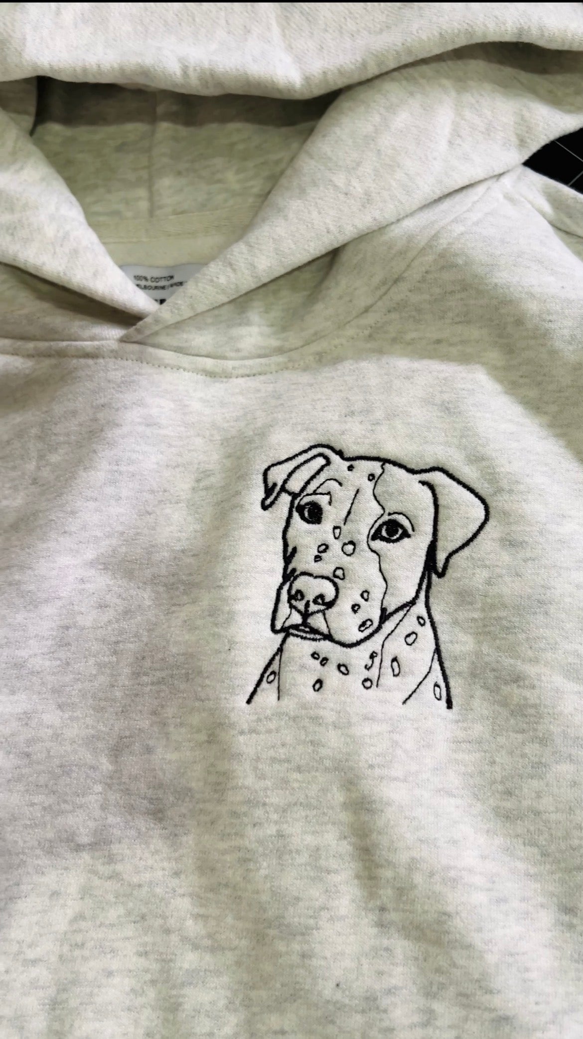 A close-up of a light heather gray hoodie featuring a black line art graphic of a spotted dog on the chest.